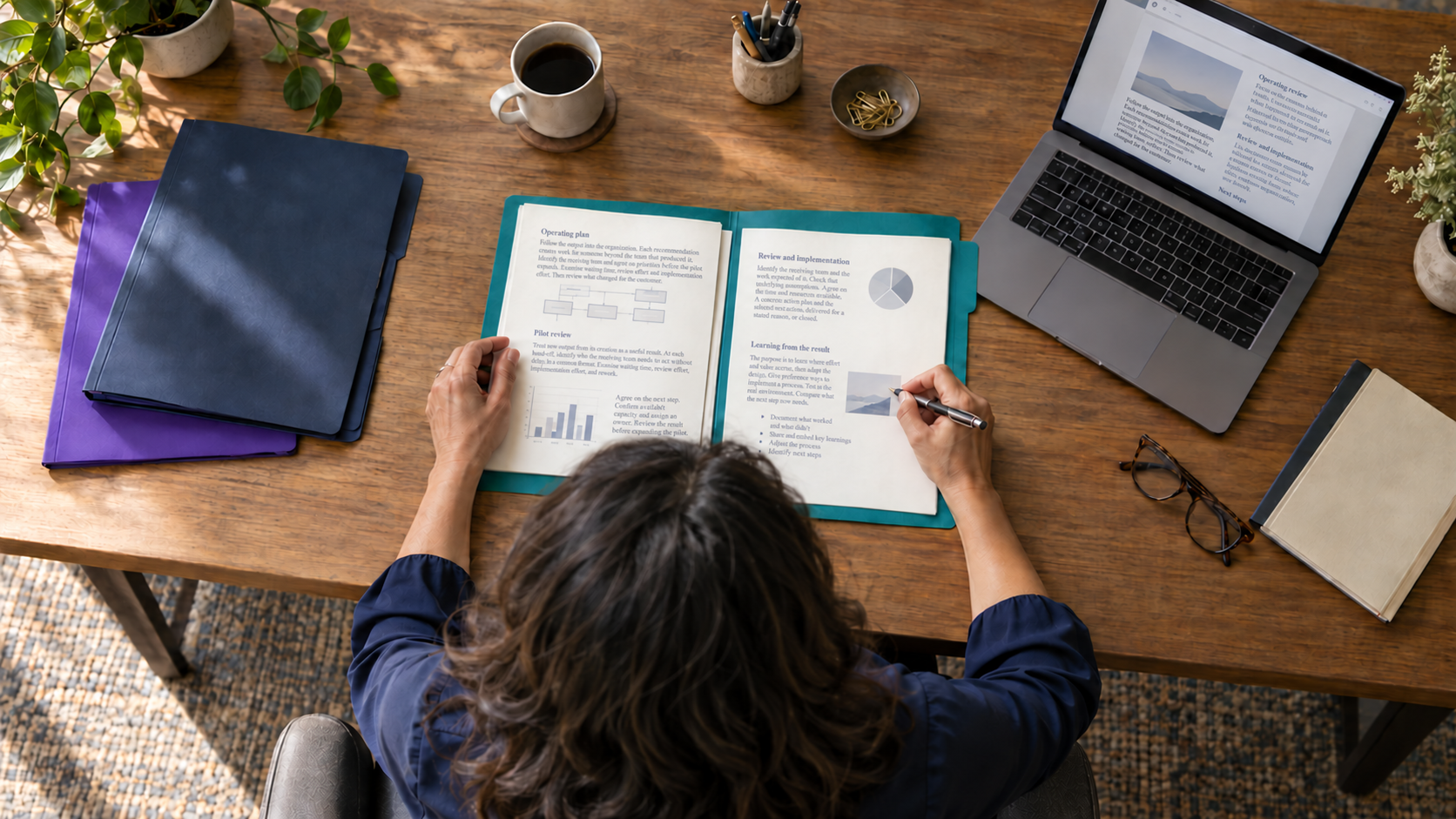 An overhead view of a person reviewing an operating plan in an open teal folder at a wooden desk, beside a laptop, notebooks, and coffee.